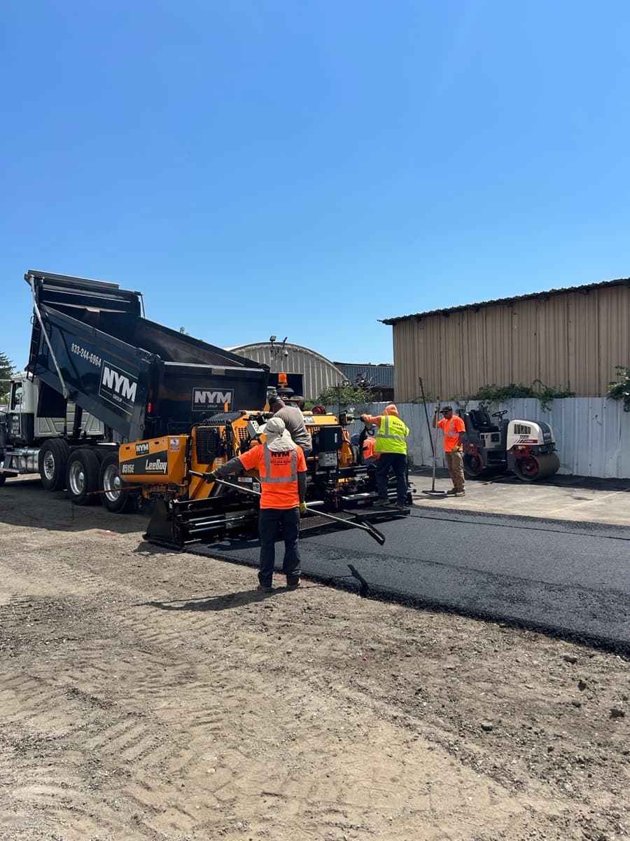 NYM crew in company hi-vis laying fresh asphalt with an NYM dump truck and paver